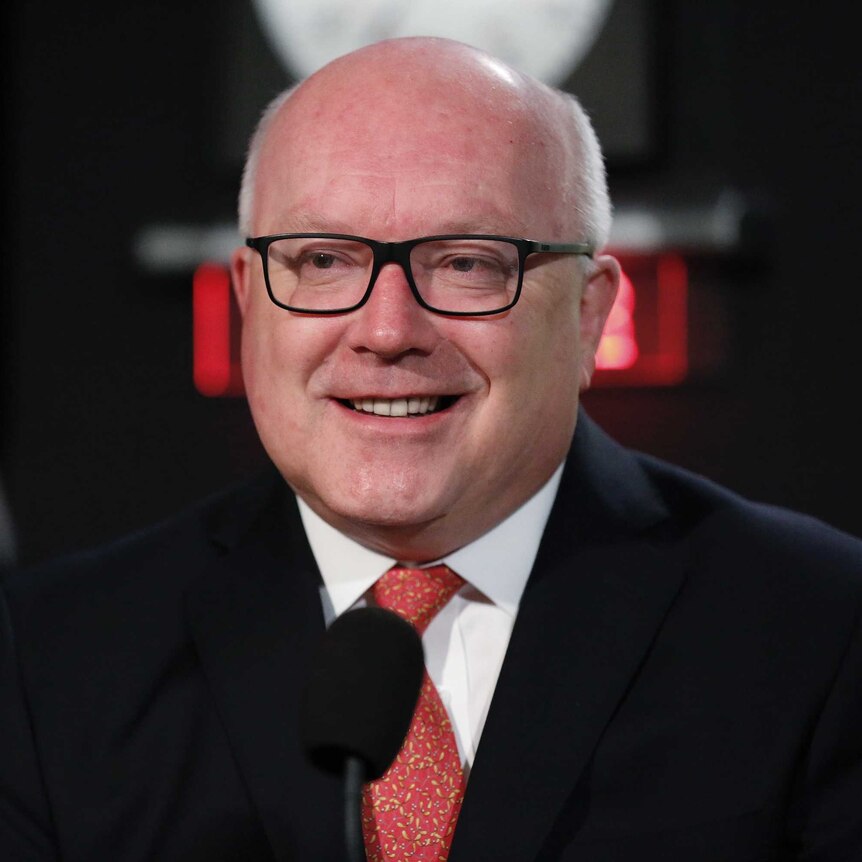 George Brandis smiles, with tears in his eyes, smiles just off camera. Behind him the ABC News logo and a clock are visible