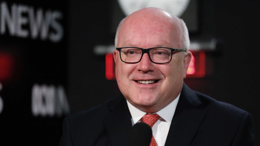 George Brandis smiles, with tears in his eyes, smiles just off camera. Behind him the ABC News logo and a clock are visible