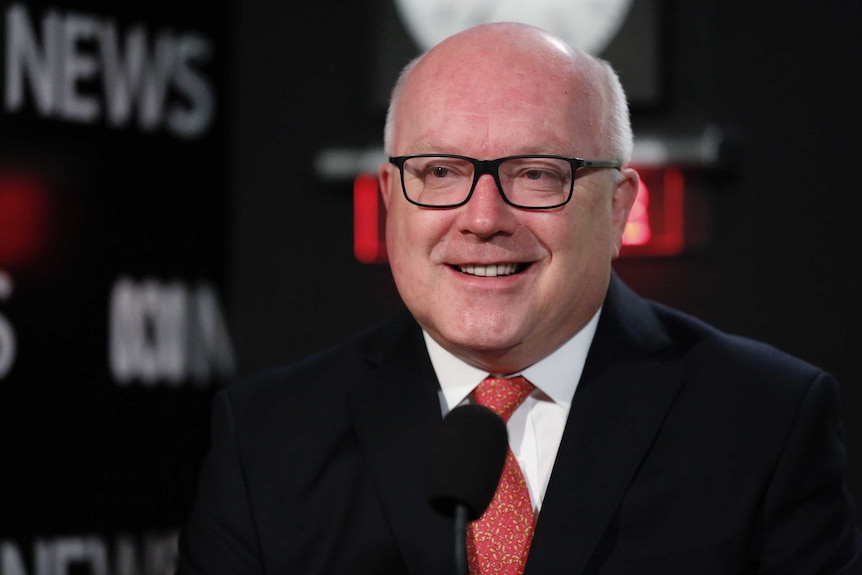 George Brandis smiles, with tears in his eyes, smiles just off camera. Behind him the ABC News logo and a clock are visible