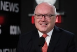 George Brandis smiles, with tears in his eyes, smiles just off camera. Behind him the ABC News logo and a clock are visible
