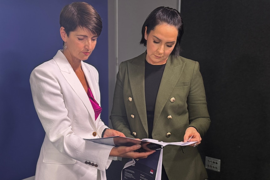 Two older women dressed in formal suits looking down at a report, both with very solemn looks on their face.