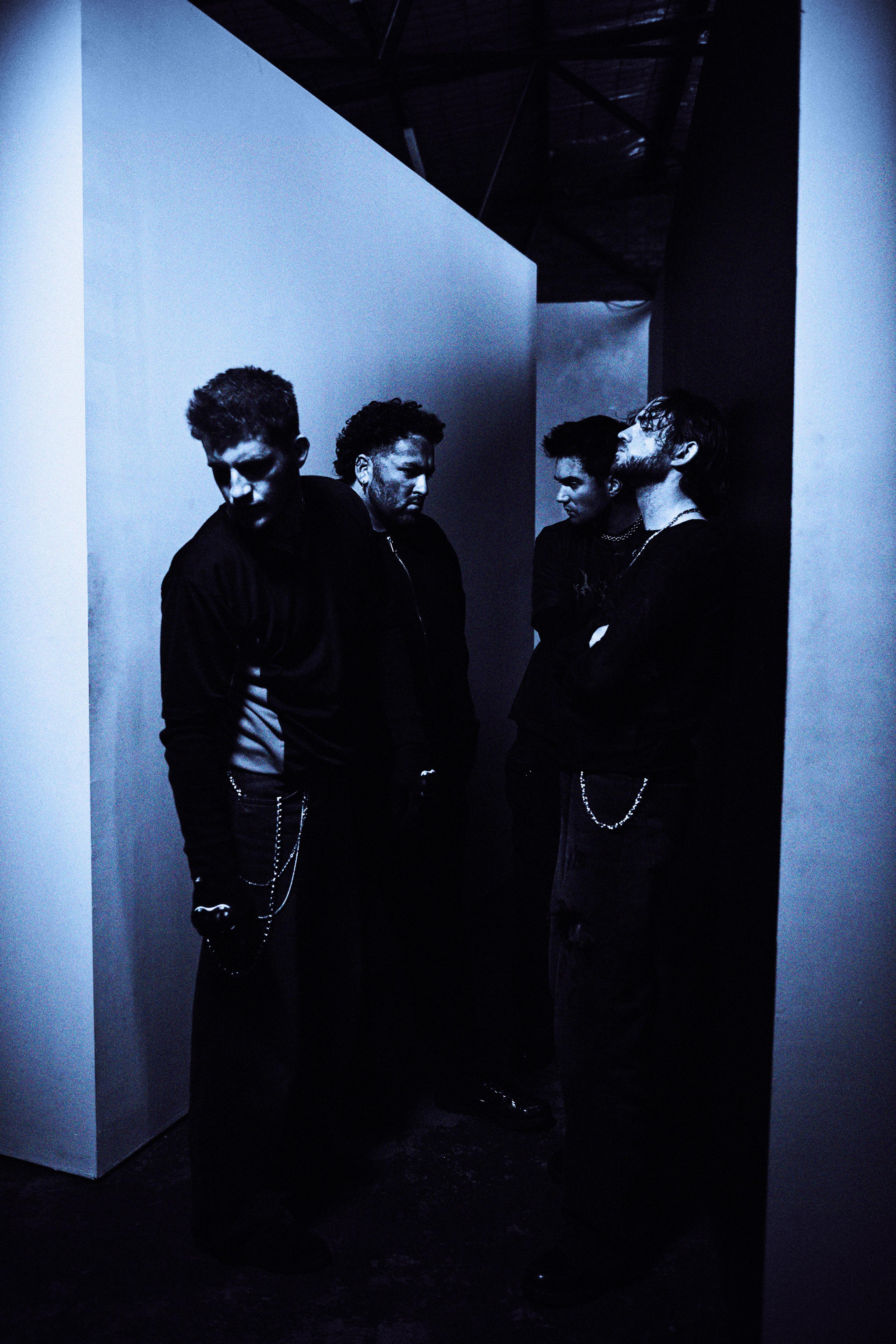 Four men are in a hallway, looking dramatic under hazy dark blue lighting. It almost looks like a film negative.