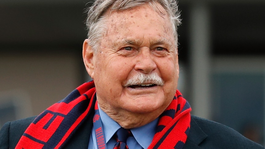 Ron Barassi smiles wearing a suit and a Melbourne Football Club scarf and lanyard