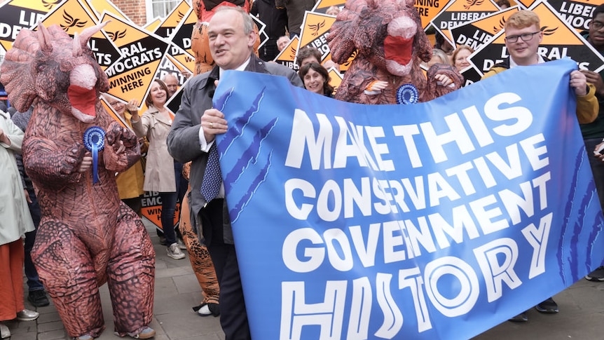Lib Dem leader greeted by a dinosaur costume and a sign saying "make this conservative government history"