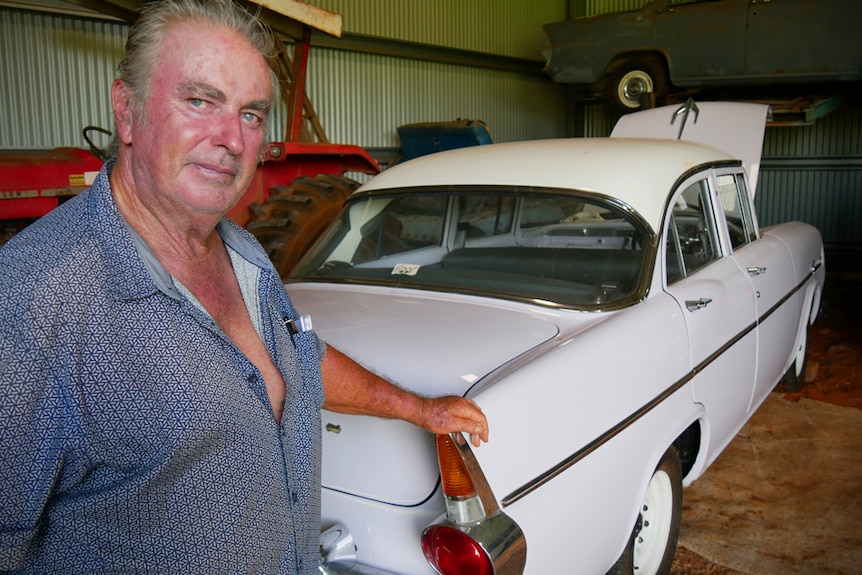 A man in front of his white holden in a shed, with another on a hoist