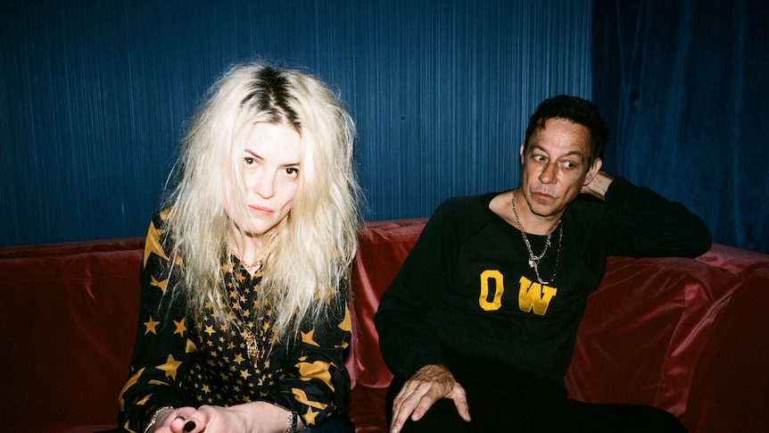 Alison Mosshart and Jamie Hince of the band The Kills sitting on a red velvet couch against a blue velvet curtain