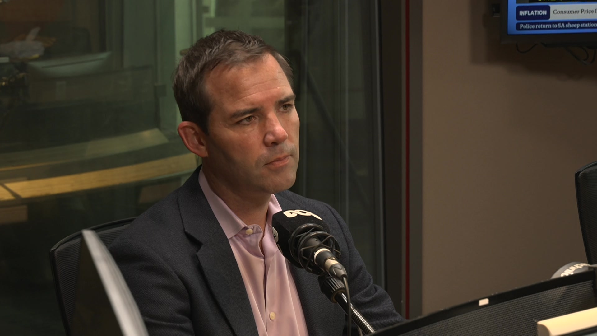 A man with brown hair in a grey blazer and pink collared shirt sits behind a microphone in a radio studio.