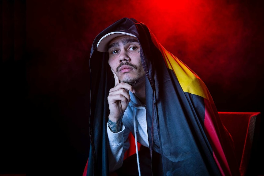Colour portrait of rapper Kobie Dee sitting with Aboriginal flag draped on him, in front of black background and some red light.