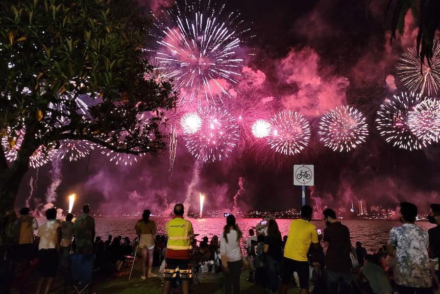 People line the Swan River at Langley Park watching as pink and red fireworks explode in the night sky.