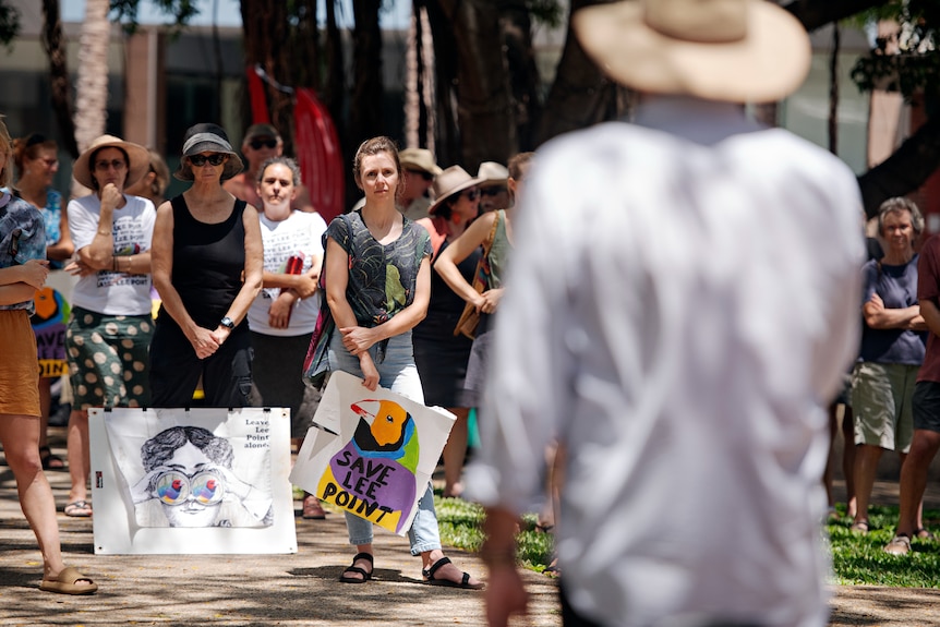 A crowd of people with protests at their feet standing outside and listening to a man speaking.