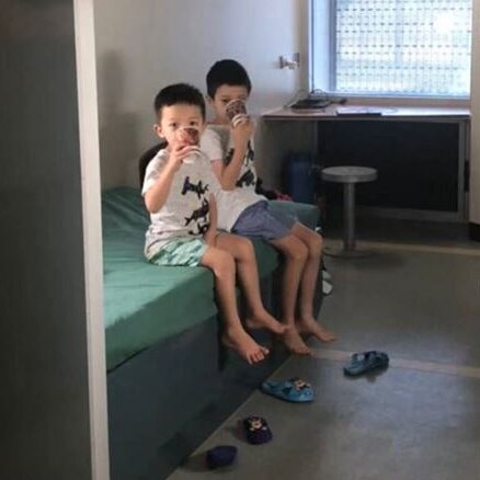 Two children sit on a bed and drink out of cups, their shoes kicked off.