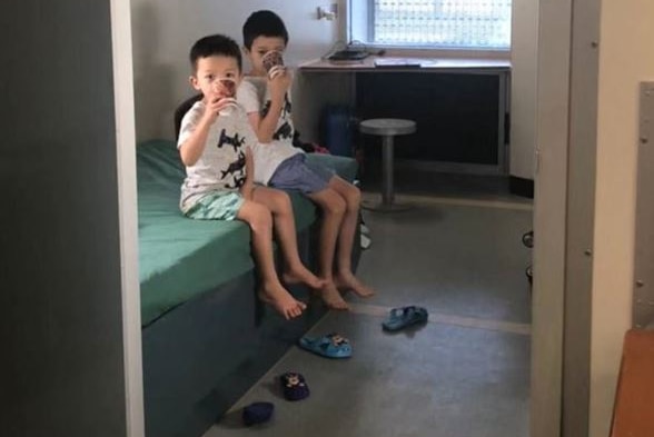 Two children sit on a bed and drink out of cups, their shoes kicked off.