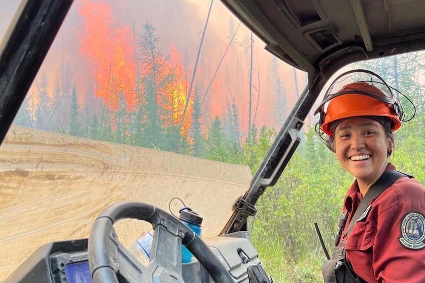 A young female firefighter sits in a golf cart and smiles at the camera. Fire is behind her.