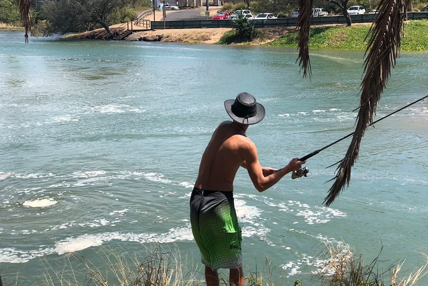 A shirtless man stands near the water's edge as he winds up to cast a fishing line.