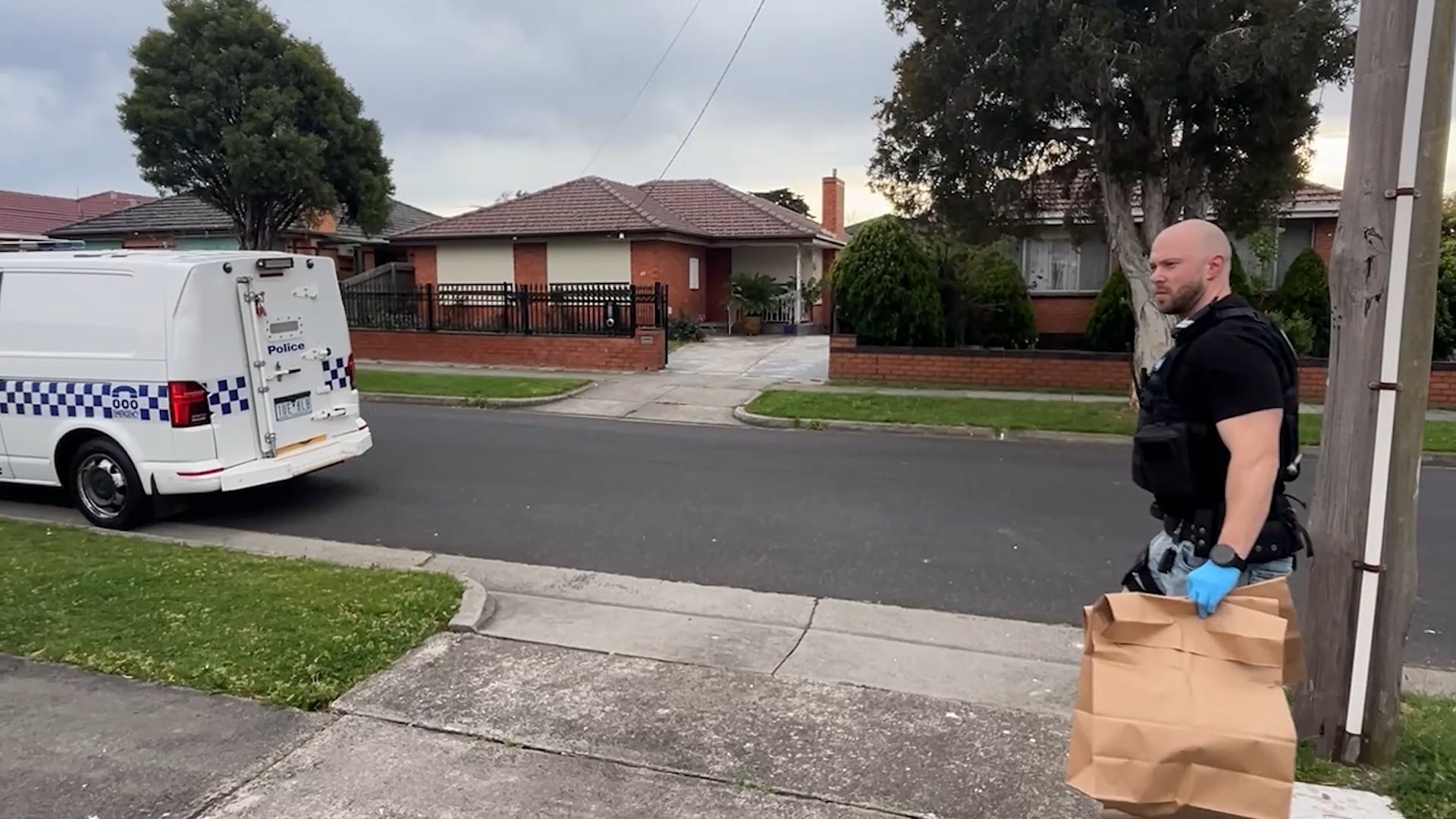 A bald man in a black shirt wearing blue gloves carries a brown paper bag towards a police van.