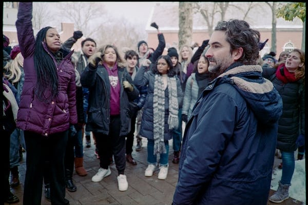 A man in a warm jacket stands in front of a group of angry students.