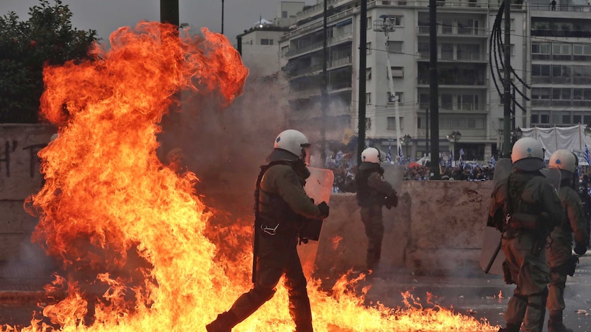 A Greek police officer walks through a bright orange fireball from a molotov cocktail explosion as three colleagues run ahead