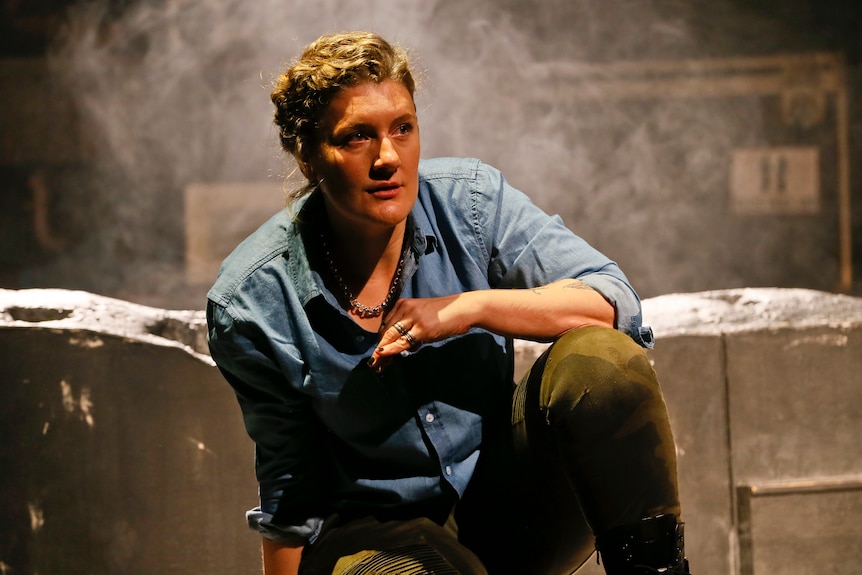 Woman with curly blonde hair wears blue denim work shirt and kneels onstage looking off into distance thoughtfully.
