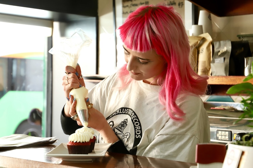 A woman withy bright pink hair pipes frosting onto cupcakes.