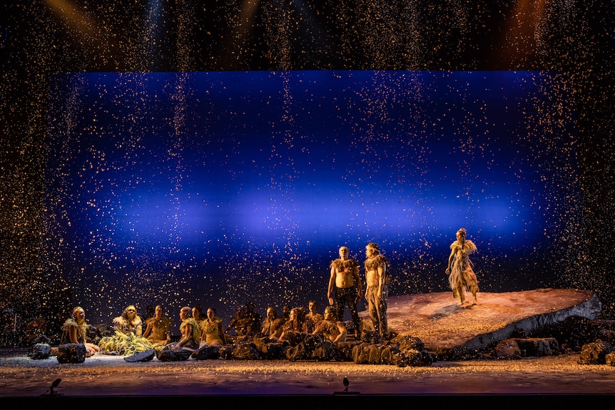 A group of Indigenous performers are mostly seated, with a couple standing on rocks, as a golden confetti-like substance falls