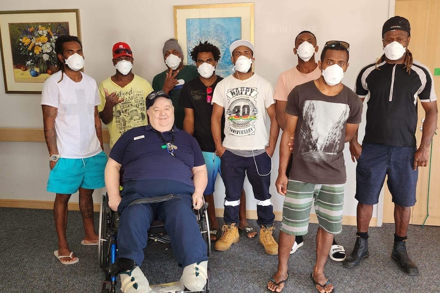 A group of nine men in a corridor wear masks. One man in wheelchair smiles to camera. 