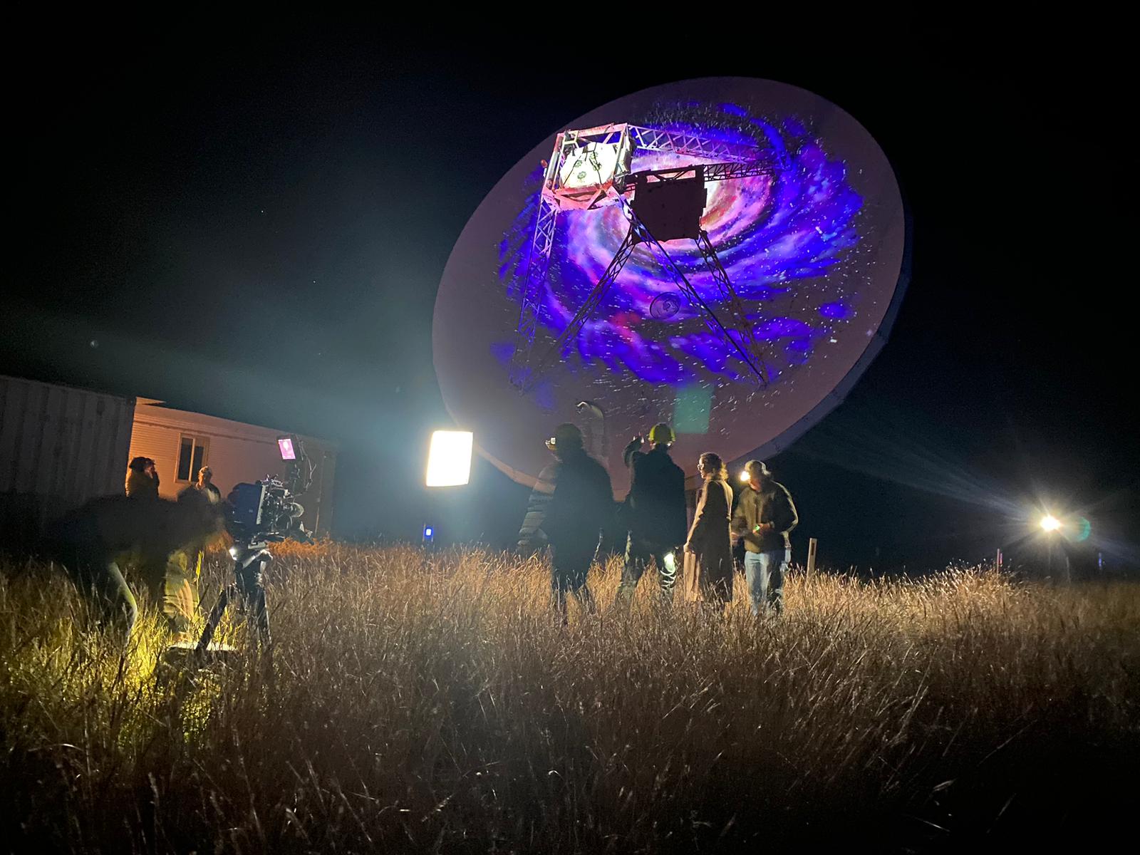 Night shot of crew filming telescope with purple light on dish.