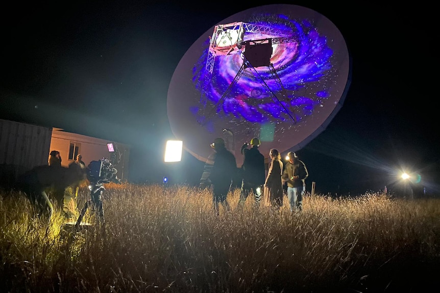Night shot of crew filming telescope with purple light on dish.