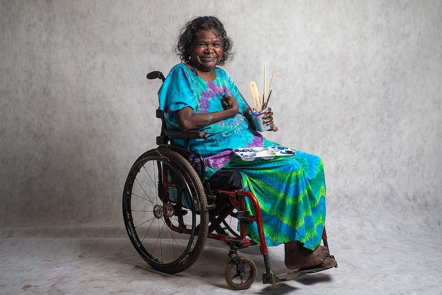 The artist Dhambit Mununggurr in bright tie-dyed top and skirt, sitting in wheelchair smiling, holding jar with brushes.