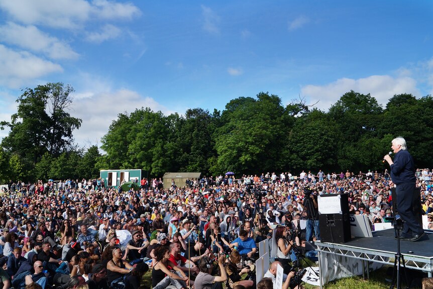 British author and commentator David Icke stands on a raised outdoor stage addressing a large crowd gathered in a field.