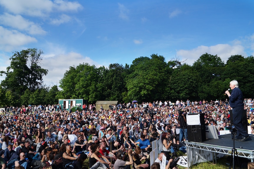 British author and commentator David Icke stands on a raised outdoor stage addressing a large crowd gathered in a field.