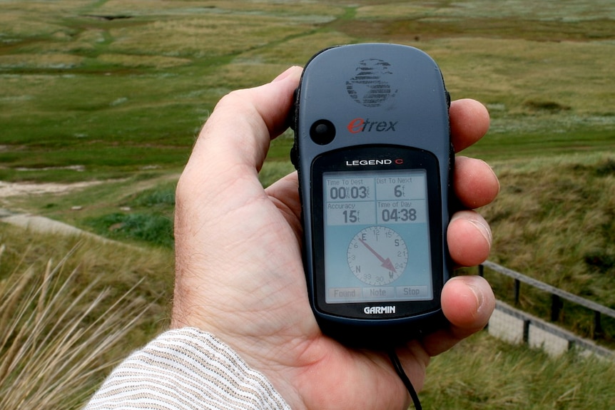 A grey GPS receiver held in a hand, with green fields in the background