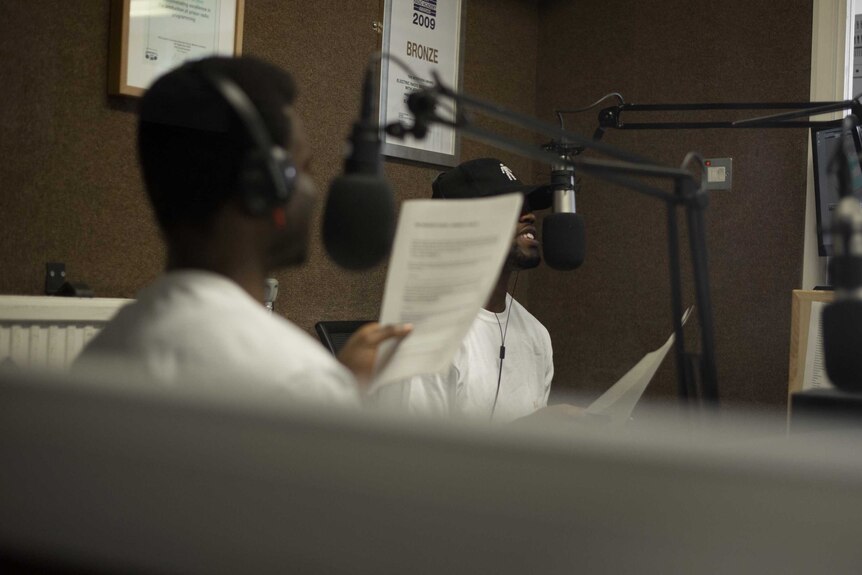 Two men holding paper and wearing headphones speak into radio microphones in a radio studio.