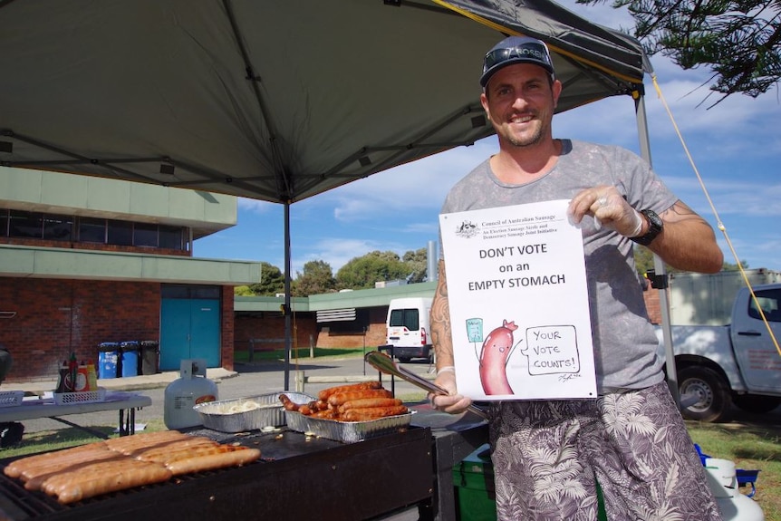 A man holds a democracy sausage poster standing next to a barbecue under a canopy.
