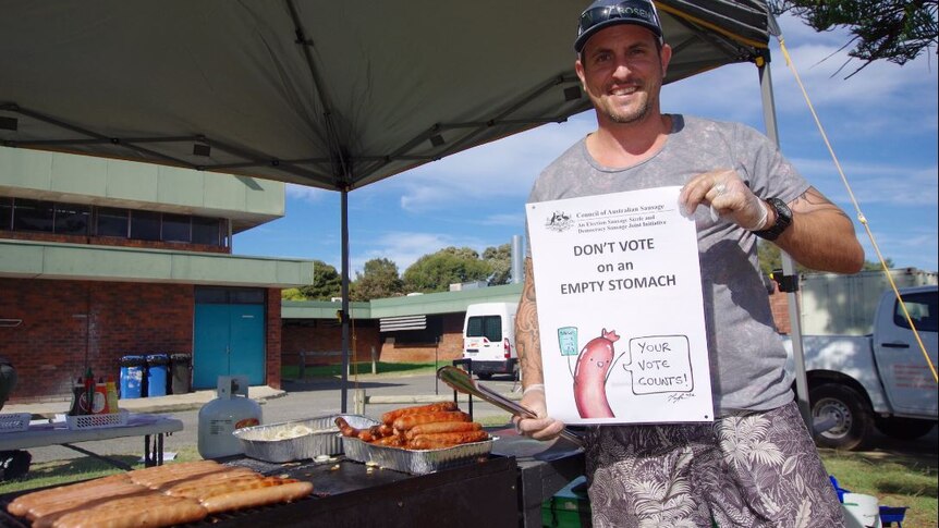 A man holds a democracy sausage poster standing next to a barbecue under a canopy.