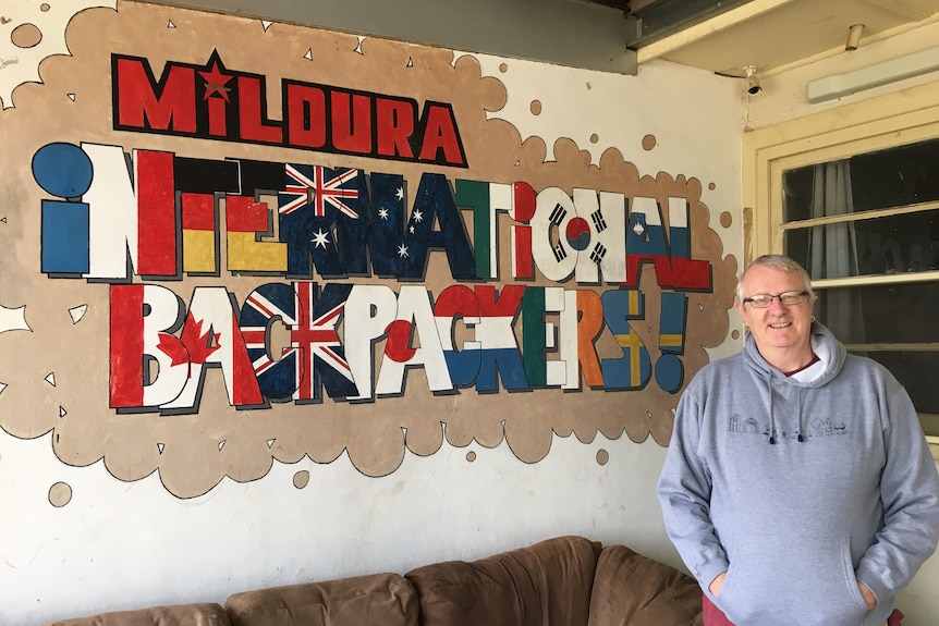 A man stands beside a mural that says "Mildura International Backpackers" and features the flags of different countries.