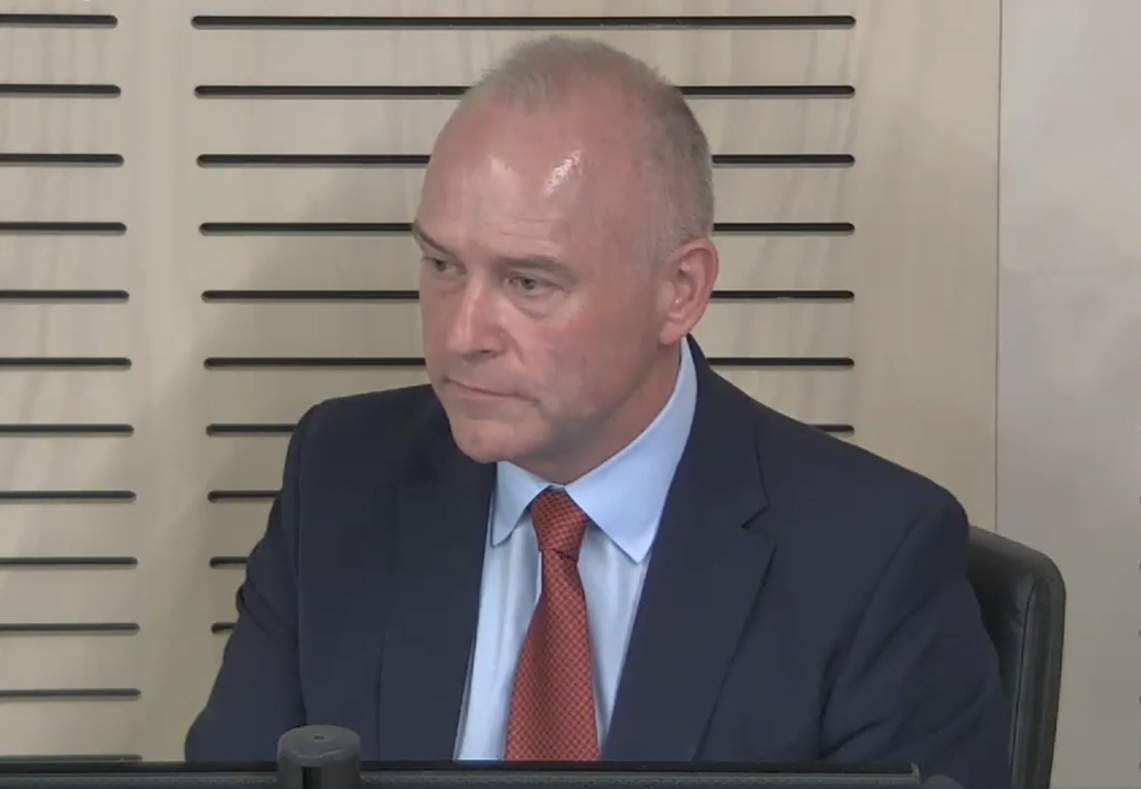 Dallas McInerney staring straight with no emotion, sitting in a commission box, wearing a suit and tie.