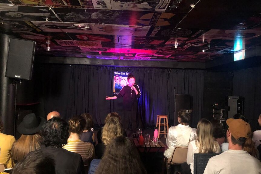 A woman stands on a stage in a small venue in front of a seated crowd doing stand up comedy