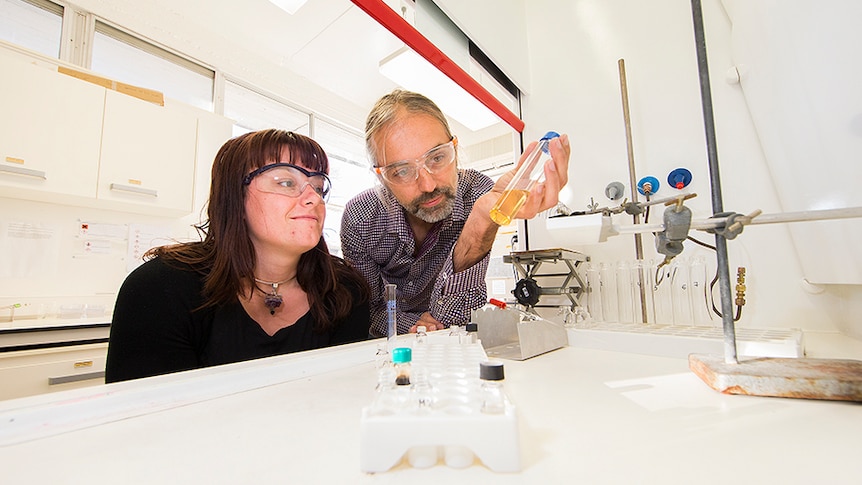 A man and a woman inside a lab, wearing safety glasses and looking at a container