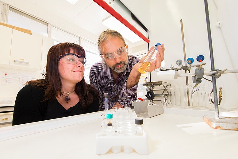 A man and a woman inside a lab, wearing safety glasses and looking at a container