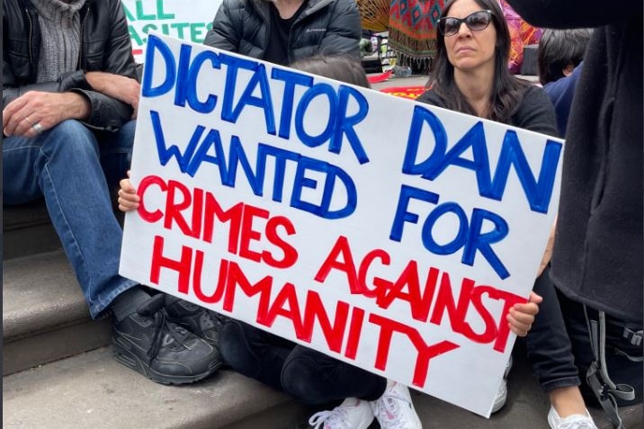 Protesters sitting on the steps of Victoria's parliament with a sign reading 'DICTATOR DAN WANTED FOR CRIMES AGAINST HUMANITY'.