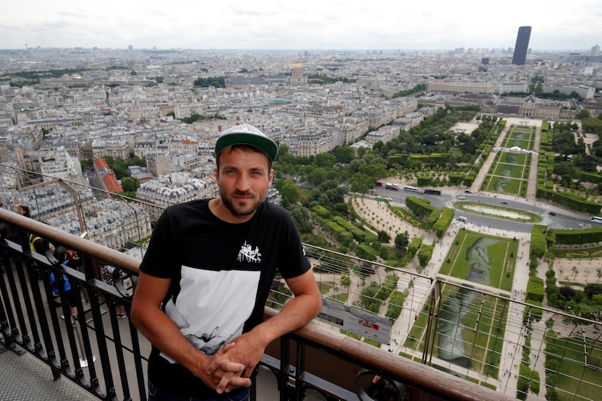El artista Saype posa en la Torre Eiffel frente a una obra de arte gigante pintada con aerosol de manos entrelazadas en los jardines de París.
