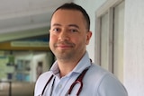 A smiling man with a stethoscope around his neck standing outside a medical clinic building. 