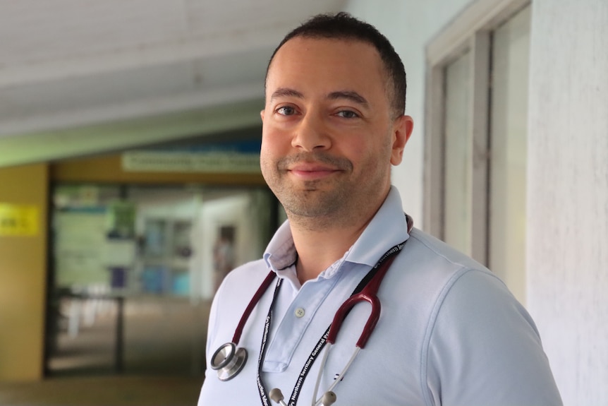 A smiling man with a stethoscope around his neck standing outside a medical clinic building. 