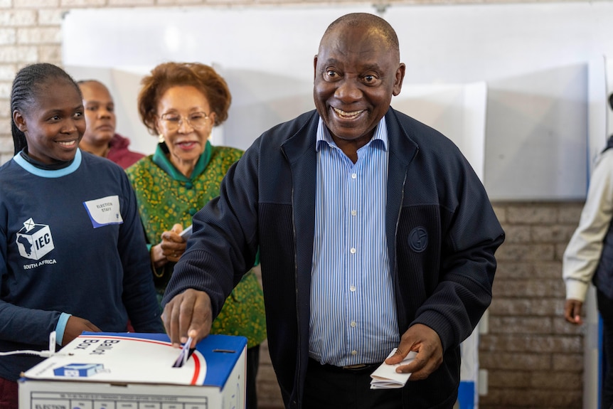 A man in a blue shirt and jacket places a paper ballot into a cardboard box smiling at the camera