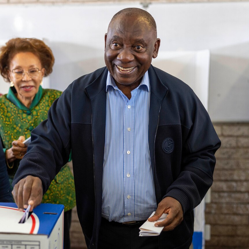A man in a blue shirt and jacket places a paper ballot into a cardboard box smiling at the camera