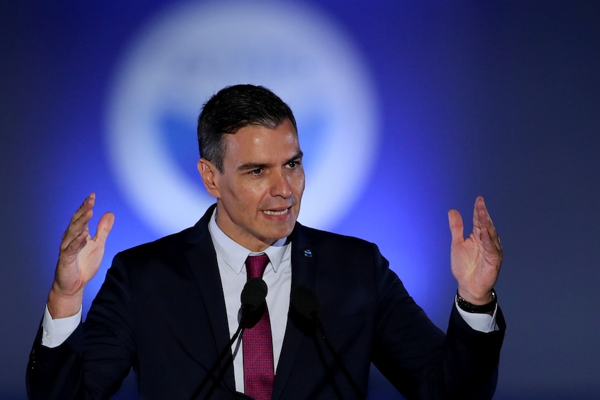 Man in a black suit with red tie speaking with his hands in the air.