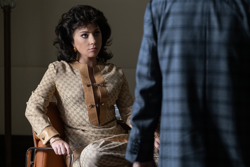 Italian woman in luxury two-piece checkered tan outfit with 60s style black wavy hair and dark eyeliner sits looking displeased.