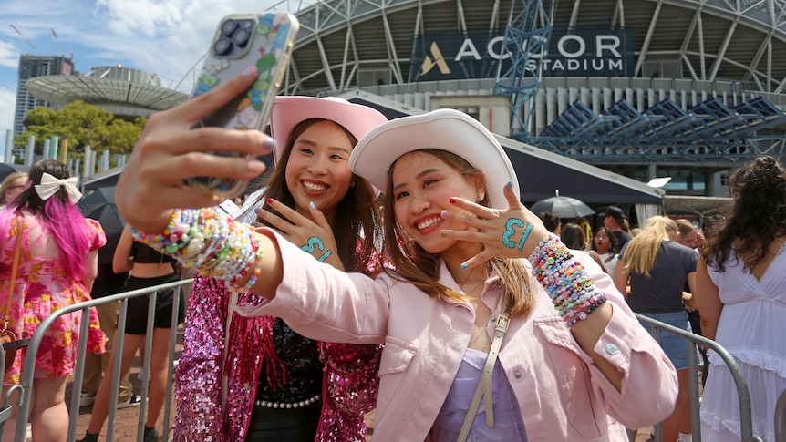 Two girls smile and pose for a selfie outside the stadium. They're both wearing pink cowgirl hats and friendship bracelets
