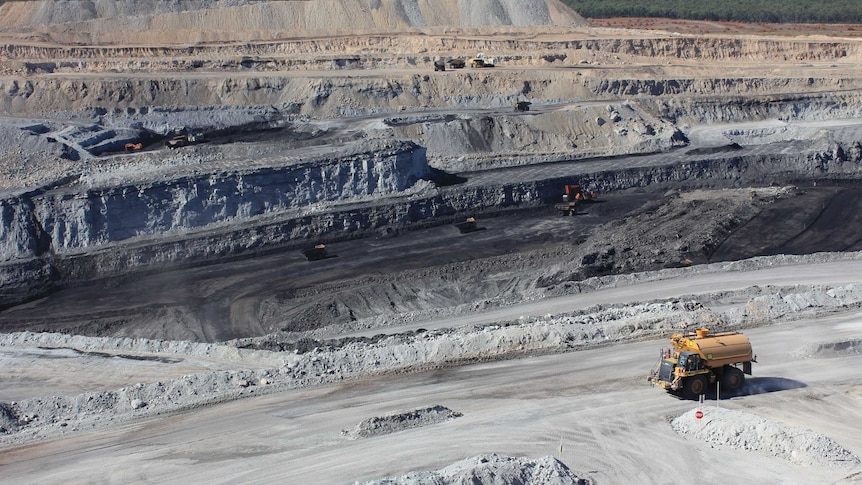 An overhead view of an open cut coal mine, with large yellow mining machine driving across the grey gravel of the mine.