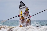 A surfboat leaps vertically out of rough surf with four women at the oars